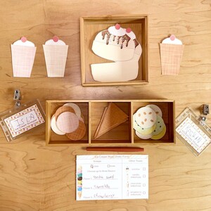 May include: A set of paper ice cream shop play items including three paper cups, a paper sundae, circular paper scoops, triangular paper cones, two plastic name badges labeled Isla and Keegan, and an Ice Cream Shop Order Form paper sheet.