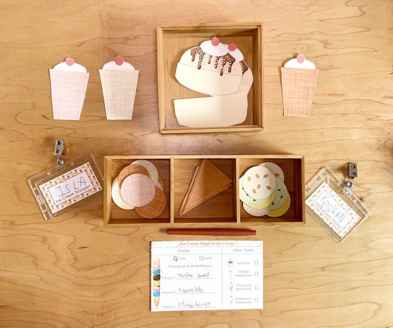 May include: A set of paper ice cream shop play items including three paper cups, a paper sundae, circular paper scoops, triangular paper cones, two plastic name badges labeled Isla and Keegan, and an Ice Cream Shop Order Form paper sheet.