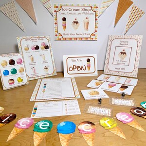 May include: An ice cream shop pretend play set featuring printed signs, menus, and order forms with illustrations of cones, sundaes, and milkshakes. Includes a banner with ice cream cone cutouts spelling out the word ice cream and name tags.