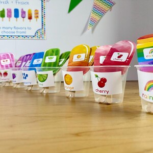 May include: A series of clear plastic cups containing colorful popsicle-shaped cutouts with wooden sticks. Each cup features a label with a fruit or rainbow illustration and corresponding text like cherry, orange, lime, and rainbow, arranged in a row.