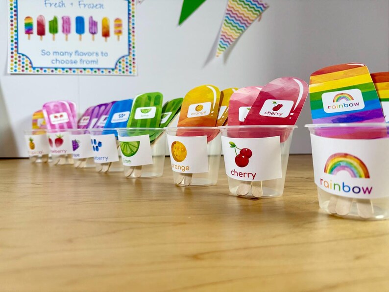 May include: A series of clear plastic cups containing colorful popsicle-shaped cutouts with wooden sticks. Each cup features a label with a fruit or rainbow illustration and corresponding text like cherry, orange, lime, and rainbow, arranged in a row.