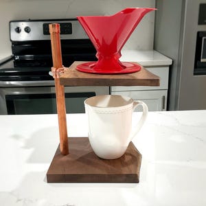 May include: A coffee pour-over stand featuring two dark wood platforms connected by a vertical copper pipe. A bright red plastic coffee dripper sits on the top platform, while a plain white ceramic mug is positioned on the bottom platform.