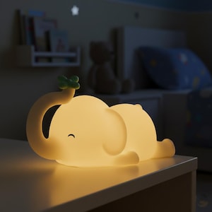 Può includere: Una luce notturna a forma di elefante giallo chiaro con un dettaglio a forma di foglia verde sulla proboscide. L'elefante è sdraiato e la luce è soffusa. La luce notturna è su una superficie bianca, con una camera da letto sullo sfondo.