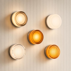 Puede incluir: Colección de cinco apliques de pared modernos con diseños circulares e iluminados. Los apliques presentan varios tonos de ámbar, blanco y vidrio transparente, sobre un respaldo de metal plateado. Las luces están montadas en una pared blanca con paneles verticales.