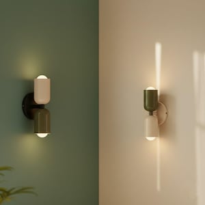 Op de afbeelding: Een moderne wandlamp met twee lampen. De bovenste lamp is lichtbeige en de onderste lamp is donker olijfgroen. De lamp is gemonteerd op een zwarte cirkelvormige basis en de lampen zijn aan.