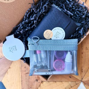 Puede incluir: Una caja de cartón abierta que contiene una bolsa con cremallera azul claro llena de botellas y recipientes pequeños. También se ve un recipiente circular blanco y pegatinas con el texto "CUTE KITS" y "TIME TO OPEN". La caja está llena de papel triturado negro.