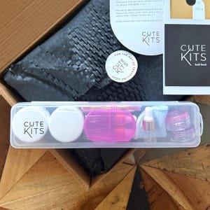 以下が含まれることがあります： 「CUTE KITS」の文字が入ったトラベルキット。白い小さな容器、ピンクのスプレーボトル、小さな透明な瓶、スポイト付きの小さなボトルが含まれています。キットは透明なプラスチックケースに入っています。