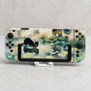 Può includere: Una console di gioco portatile con una custodia protettiva trasparente con un design stampato di un gatto nero su una ninfea in uno stagno con foglie verdi e fiori bianchi. La console ha joystick, pulsanti e un supporto a cornice di colore nero.