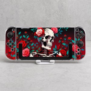 Puede incluir: Una consola de juegos portátil con una funda decorativa que presenta una ilustración central de un cráneo humano adornado con rosas rojas y hojas verde azulado sobre un fondo rojo oscuro. El dispositivo incluye joysticks y botones negros.