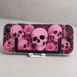 Puede incluir: Una consola de juegos portátil con una funda de color rosa y magenta oscuro con un patrón de calaveras humanas y flores. El dispositivo tiene una carcasa protectora transparente y está montado sobre un soporte de plástico blanco con botones negros.
