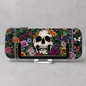 May include: A handheld gaming console featuring a clear protective case with a printed design of a central human skull surrounded by colorful flowers, green leaves, a monarch butterfly, a purple butterfly, and a small bee on a dark background.