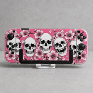 May include: A handheld gaming console skin featuring a vibrant pink background decorated with a repeating pattern of white human skulls and delicate pink cherry blossoms. The design covers the front face of the device, including the controller areas.