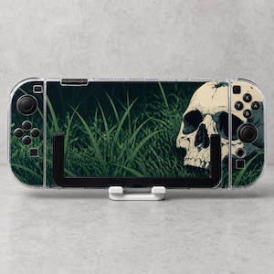 May include: A handheld gaming console featuring a clear protective case and a skin decal. The decal displays a detailed illustration of a human skull resting in dark green grass. The console includes black joysticks, directional buttons, and action buttons.
