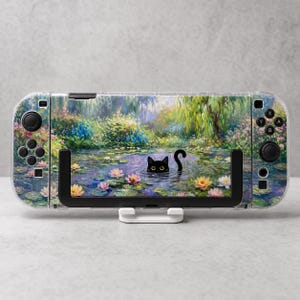 Puede incluir: Una consola de juegos portátil con una funda protectora que muestra una pintura impresionista de un estanque lleno de nenúfares coloridos y un gato negro asomándose desde el agua. El dispositivo tiene botones y joysticks negros.