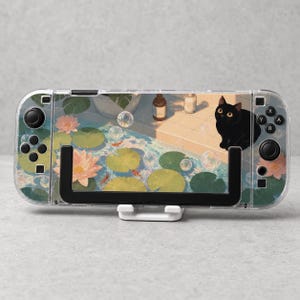 May include: A clear protective case for a handheld gaming console featuring a printed illustration of a black cat sitting by a pond with pink lotus flowers, green lily pads, orange koi fish, and floating bubbles on a tiled surface.
