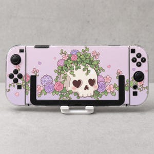 May include: A handheld gaming console featuring a light purple skin with a central illustration of a white skull with heart-shaped eye sockets, surrounded by green ivy vines, pink and purple flowers, and small scattered heart shapes.