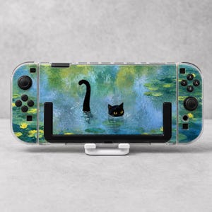 Pode incluir: Uma consola de jogos portátil com uma capa que apresenta uma ilustração de um gato preto a nadar num lago com água azul, nenúfares verdes e flores amarelas. A consola tem botões, joysticks e uma moldura de cor preta.