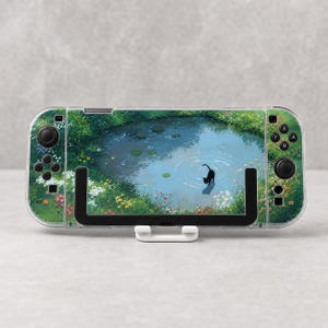 May include: A handheld gaming console featuring a clear protective case and a decorative skin depicting a black cat standing in a blue pond surrounded by green grass and colourful wildflowers. The device is mounted on a white plastic stand.