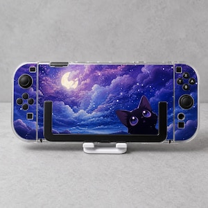 May include: A handheld gaming console featuring a skin with a dark purple night sky, a bright yellow full moon, and a black cat with large purple eyes. The device has black buttons, joysticks, and a clear protective case, held by a white stand.