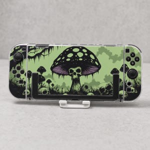 May include: A handheld gaming console featuring a light green skin with a black graphic print of a large mushroom with a skull base, surrounded by smaller mushrooms and skulls. The device is held in a black plastic stand with a white base.