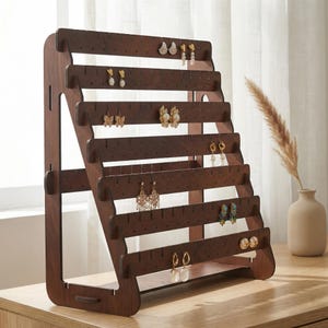 May include: A dark brown wooden earring display stand with multiple horizontal bars, each with small holes for hanging earrings. Various earrings of different styles and colors are displayed on the stand. The stand is on a light-colored wooden surface.
