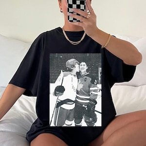 May include: Black t-shirt featuring a black and white image of two hockey players. One player is kissing the other on the cheek. The players are wearing hockey jerseys and helmets. The image is centered on the shirt.