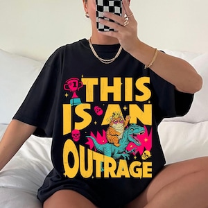 May include: Black t-shirt with a colorful graphic design. The design features a cat wearing a crown riding a dinosaur, with the words "THIS IS AN OUTRAGE" in yellow. Additional elements include skulls and a trophy.
