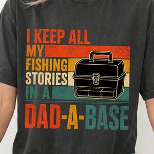 Könnte beinhalten: Ein anthrazitfarbenes T-Shirt mit einer Grafik aus horizontalen Streifen in Orange, Gelb und Blaugrün. Der Text lautet I KEEP ALL MY FISHING STORIES IN A DAD-A-BASE, ergänzt durch eine schwarze Strichzeichnung einer klassischen Angelkiste.
