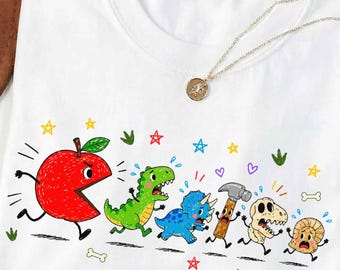 May include: A white crew neck t-shirt featuring a colorful graphic of a large red apple chasing a green dinosaur, a blue triceratops, a hammer, a skull, and a small round object. The design includes small stars, hearts, and bone shapes.