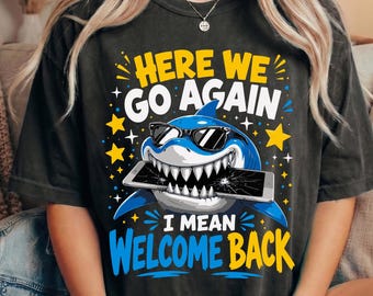 May include: A charcoal gray short-sleeve t-shirt featuring a graphic of a blue shark wearing black sunglasses and biting a cracked smartphone. The text reads HERE WE GO AGAIN I MEAN WELCOME BACK in yellow, white, and blue block lettering.