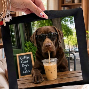 May include: A square black frame featuring a photograph of a brown Labrador dog wearing black sunglasses. The dog is sitting at a wooden table with an iced coffee in a clear plastic cup. A small chalkboard sign in the background reads Coffee Good Days & Labs.