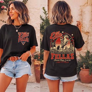 May include: A black short-sleeve t-shirt featuring a small graphic on the front chest with the text Ella Fellas. The back displays a large graphic of a cowboy on a horse with a sunset, cactus, and the text Ella Fellas Choosin Texas, I can tell.