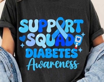 May include: A black t-shirt featuring the text "SUPPORT SQUAD DIABETES AWARENESS" in shades of blue. The design includes a blue awareness ribbon and a graphic of a blood glucose monitor with a heart icon, all rendered in a bold, bubbly font.