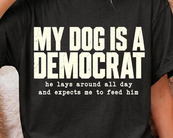May include: A black short-sleeved t-shirt featuring the white distressed-style text "MY DOG IS A DEMOCRAT" in large capital letters, followed by the smaller text "he lays around all day and expects me to feed him" centered on the chest.