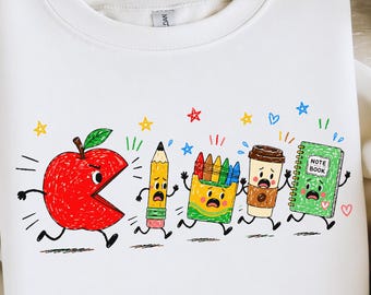 May include: A white crewneck sweatshirt featuring a graphic print of a large red apple chasing a pencil, a box of crayons, a coffee cup, and a green notebook. Each item has a cartoon face with a panicked expression and small limbs in motion.