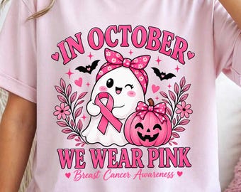May include: A light pink t-shirt featuring a graphic of a white ghost wearing a pink floral headband, holding a pink breast cancer awareness ribbon next to a pink pumpkin. The text reads IN OCTOBER WE WEAR PINK Breast Cancer Awareness.