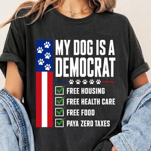 May include: A charcoal gray short-sleeve t-shirt featuring the text MY DOG IS A DEMOCRAT with a vertical American flag design, paw prints, and a checklist including Free Housing, Free Health Care, Free Food, and Paya Zero Taxes in white text.