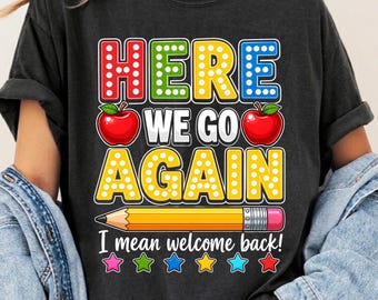 May include: A charcoal gray t-shirt featuring the text "HERE WE GO AGAIN" in colorful, dotted block letters, flanked by two red apples. Below, a yellow pencil graphic sits above the text "I mean welcome back!" and a row of six colorful stars.