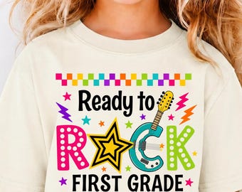 May include: A cream-colored t-shirt featuring a colorful graphic with the text Ready to ROCK FIRST GRADE. The design includes a yellow star, a teal electric guitar, and a checkered border in orange, yellow, green, and purple accents.