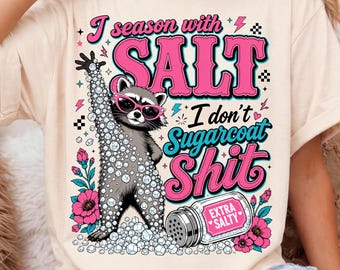May include: A cream-colored t-shirt featuring a graphic of a raccoon wearing pink sunglasses, surrounded by salt crystals and flowers. The text reads: I season with SALT I don't Sugarcoat Shit, with a salt shaker labeled Extra Salty at the bottom.
