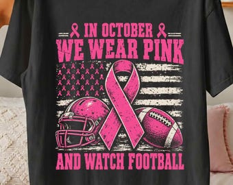 May include: A black t-shirt featuring a pink graphic with the text IN OCTOBER WE WEAR PINK AND WATCH FOOTBALL. The design includes a pink awareness ribbon, a football helmet, and a football, all set against a stylized American flag background.