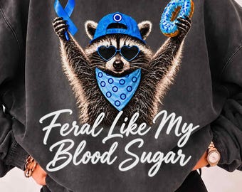 May include: A charcoal grey mineral-washed sweatshirt featuring a graphic of a raccoon wearing a blue baseball cap, heart-shaped sunglasses, and a blue bandana. The raccoon holds a blue ribbon and a blue donut. Text reads: Feral Like My Blood Sugar.