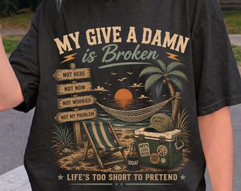 May include: A black t-shirt featuring a graphic print with the text "MY GIVE A DAMN is Broken" and "LIFE'S TOO SHORT TO PRETEND". The design includes a beach scene with a hammock, a palm tree, a wooden signpost, a beach chair, and a cooler.