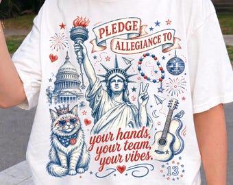 May include: A white t-shirt featuring a graphic print of the Statue of Liberty, the U.S. Capitol building, a cat wearing heart-shaped sunglasses, a guitar, and a disco ball. The text reads, "PLEDGE ALLEGIANCE TO your hands, your team, your vibes."