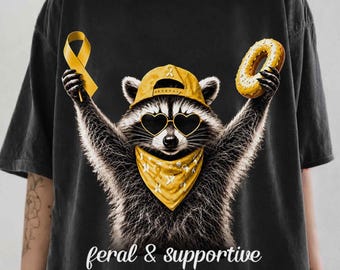 May include: A charcoal gray t-shirt featuring a graphic of a raccoon wearing a yellow baseball cap, heart-shaped sunglasses, and a yellow bandana. The raccoon holds a yellow awareness ribbon and a donut. The text reads feral & supportive.