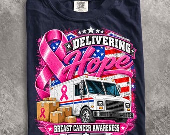 May include: A navy blue t-shirt featuring a graphic of a white delivery truck, brown cardboard boxes, and a large pink ribbon. The text reads Delivering Hope and Breast Cancer Awareness, set against a background of a stylized American flag.