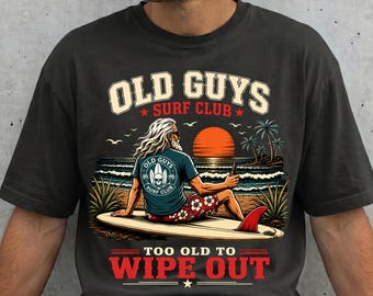 May include: A charcoal gray t-shirt featuring a graphic of a person with long white hair sitting on a surfboard at the beach. The text reads OLD GUYS SURF CLUB and TOO OLD TO WIPE OUT. The scene includes a sunset, ocean waves, and palm trees.