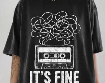 May include: A black short-sleeve t-shirt featuring a white graphic of a cassette tape with tangled tape spilling out from the top. Below the cassette tape, the text IT'S FINE is printed in a bold, white, distressed-style sans-serif font.