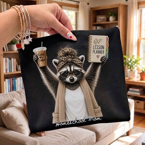 May include: A black fabric item featuring a graphic of a raccoon with a messy bun, leopard print headband, and glasses. The raccoon wears a white shirt and tan scarf, holding a coffee cup labeled Fueled by Coffee and a Lesson Planner. Text reads Homeschool Mom.