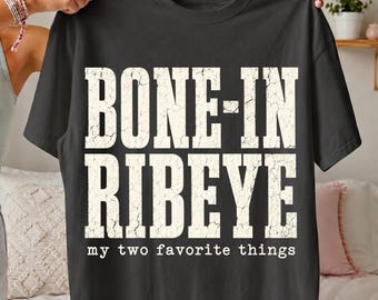May include: A black short-sleeve t-shirt featuring the text BONE-IN RIBEYE in large, distressed, off-white capital letters, with the smaller text my two favorite things printed in a simple white font directly underneath the main graphic.